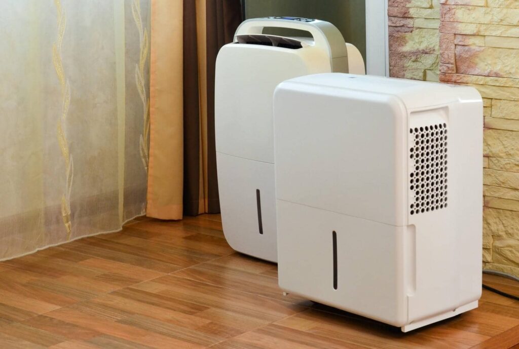 Two white dehumidifiers placed on a wooden floor near curtains and a stone wall.